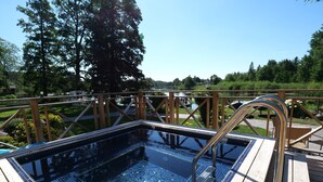A rectangular swimming pool with a tiled deck and metal handrails, surrounded by a wooden deck and metal railing, overlooking a landscaped area with trees and a body of water.