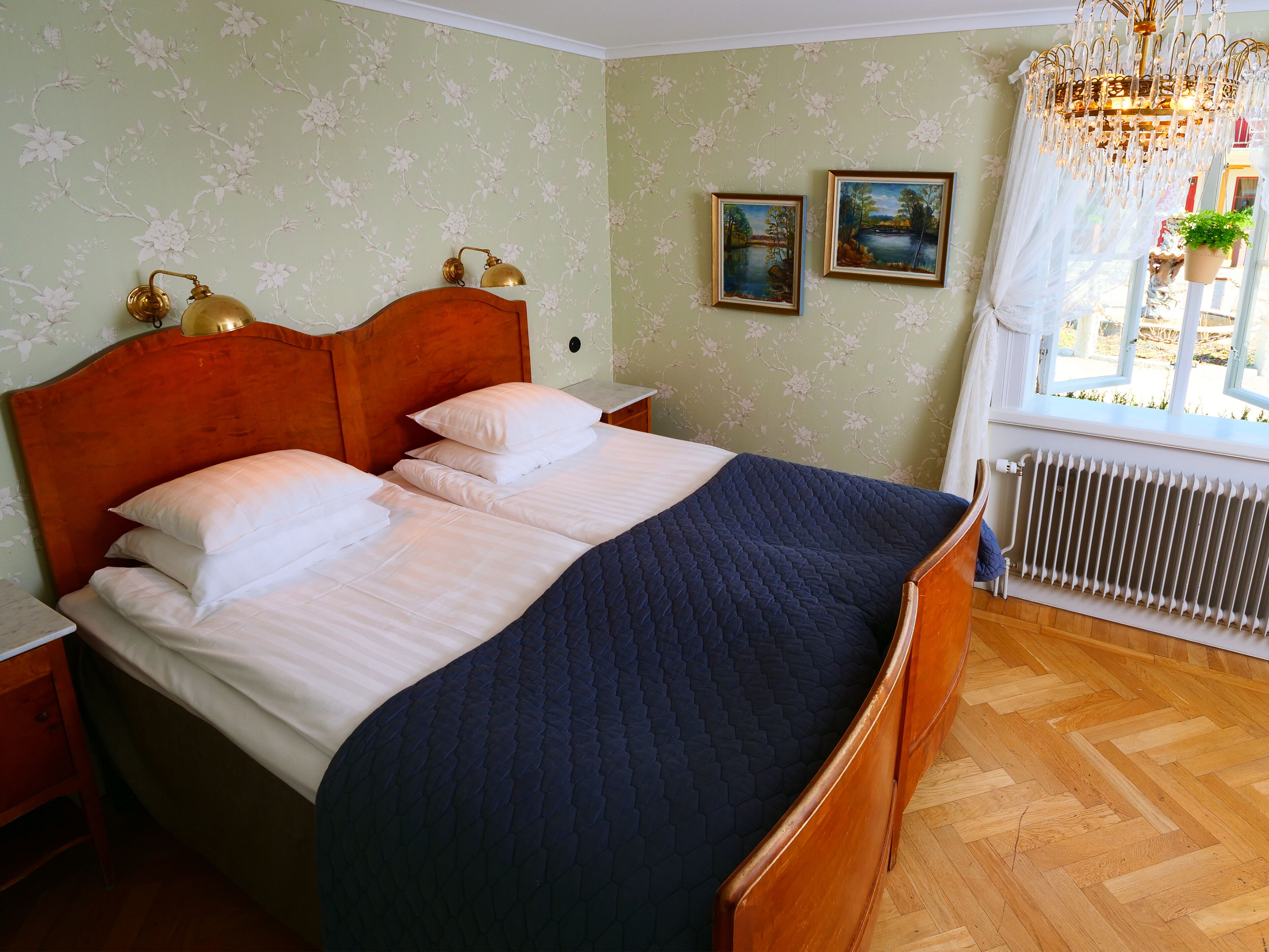 A neatly made bed with white and navy bedding, a wooden headboard, and a nightstand with a brass lamp. There are two framed paintings on the wall and a window with a view of the outdoors.