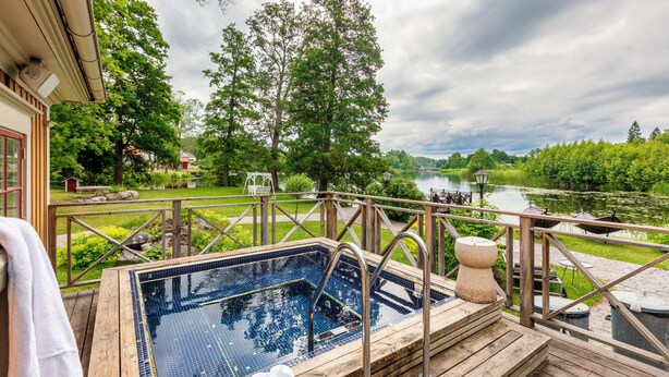 A wooden deck with a hot tub overlooking a lake, surrounded by lush greenery and trees.