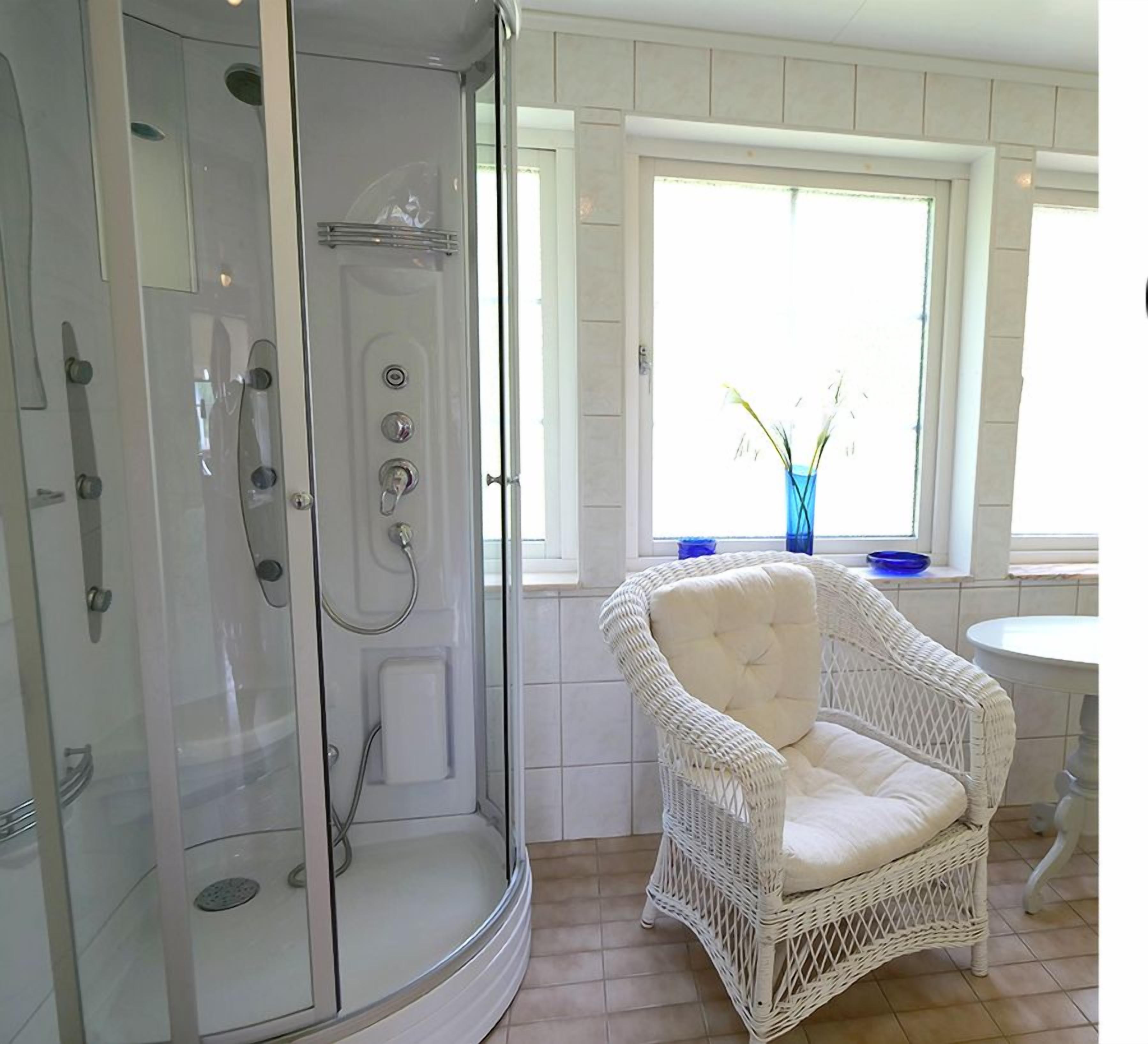 Een badkamer met een glazen douchecabine, een witte rotanstoel en een raam met een bloevas.