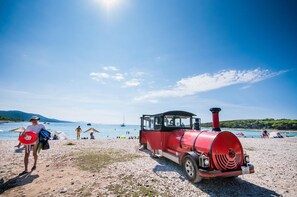 Un train rouge sur une plage, avec des personnes et des bateaux en arrière-plan.