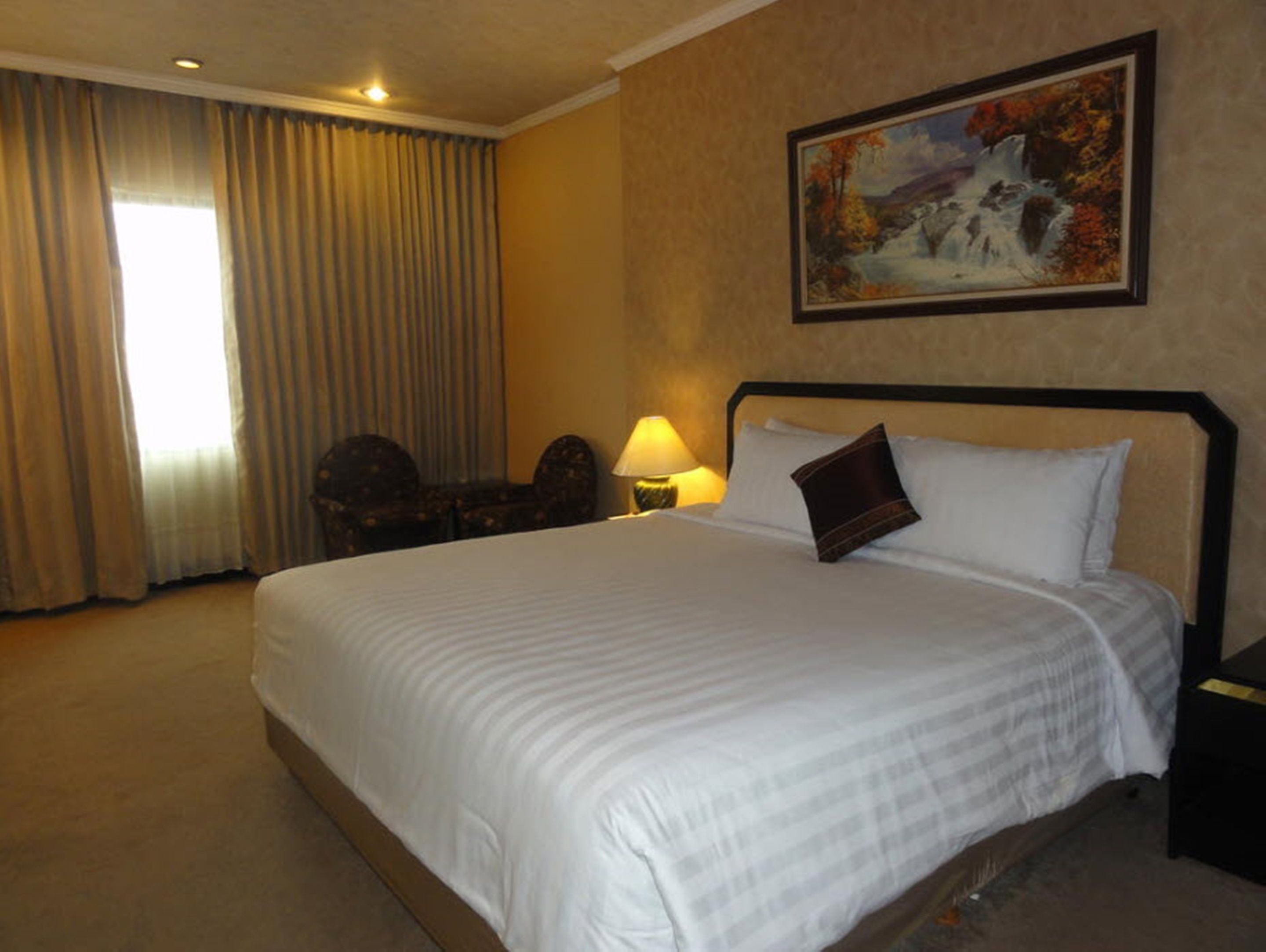 A hotel room with a large bed, a nightstand with a lamp, a chair, and a painting on the wall.