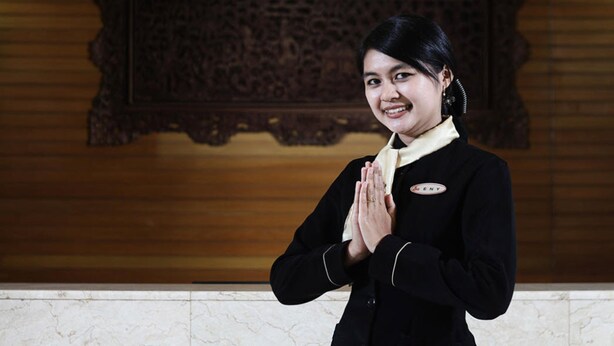 A person in a black uniform with a name tag, standing in front of a wooden wall with an ornate carving. - Regent's Park Hotel (Malang)