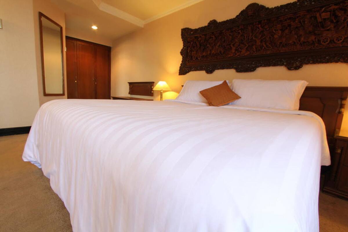 A hotel room with a large bed, a wooden headboard, a nightstand with a lamp, and a wardrobe.