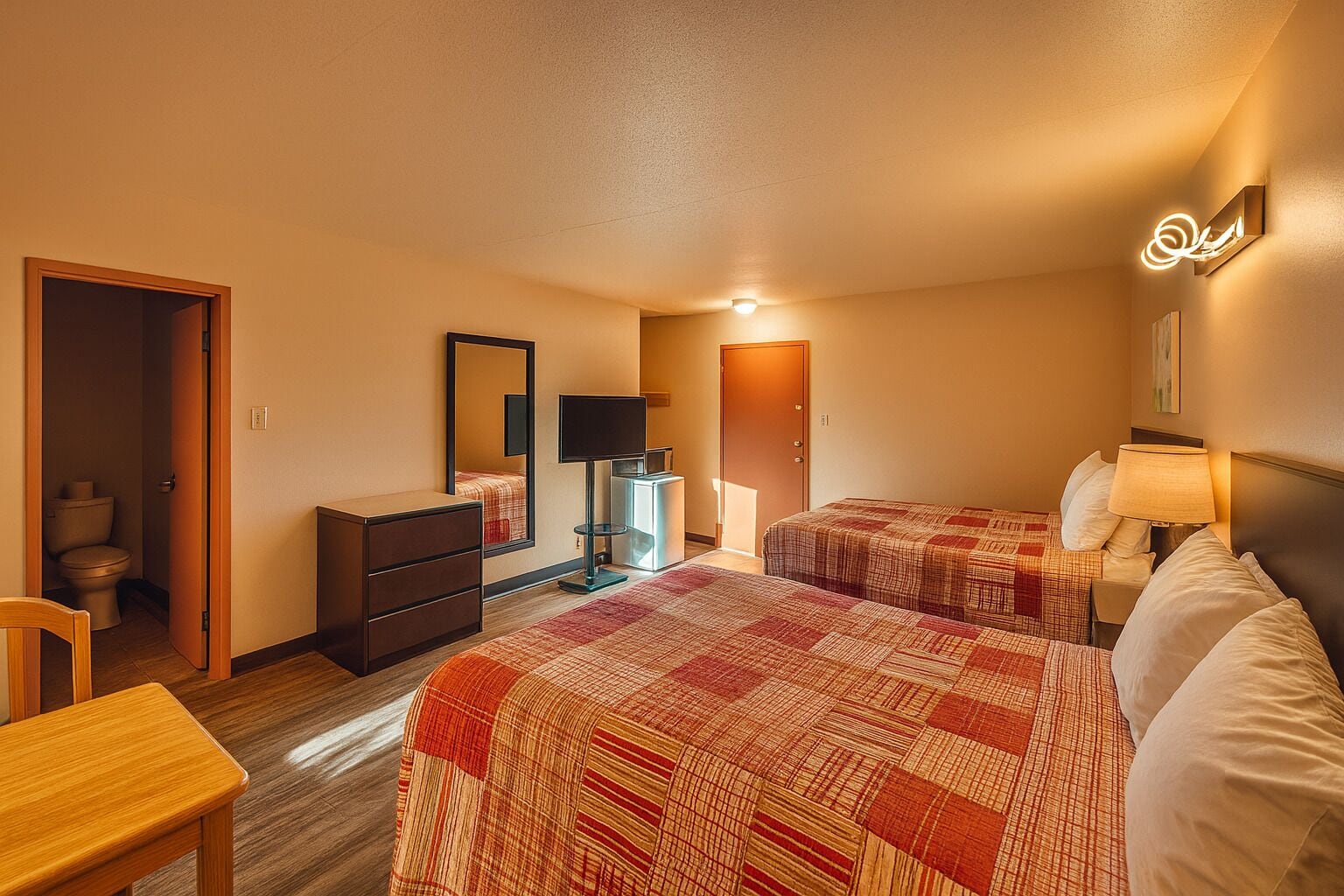 Une chambre d’hôtel avec deux lits, une commode, un bureau, un miroir, une télévision et une porte donnant sur une salle de bain.