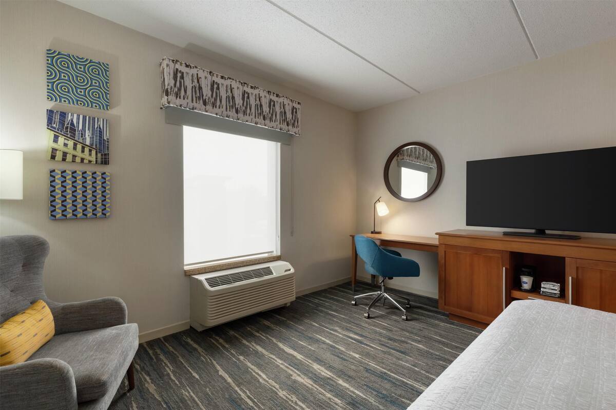 A hotel room with a large window, a desk with a chair, a television, and a bed.