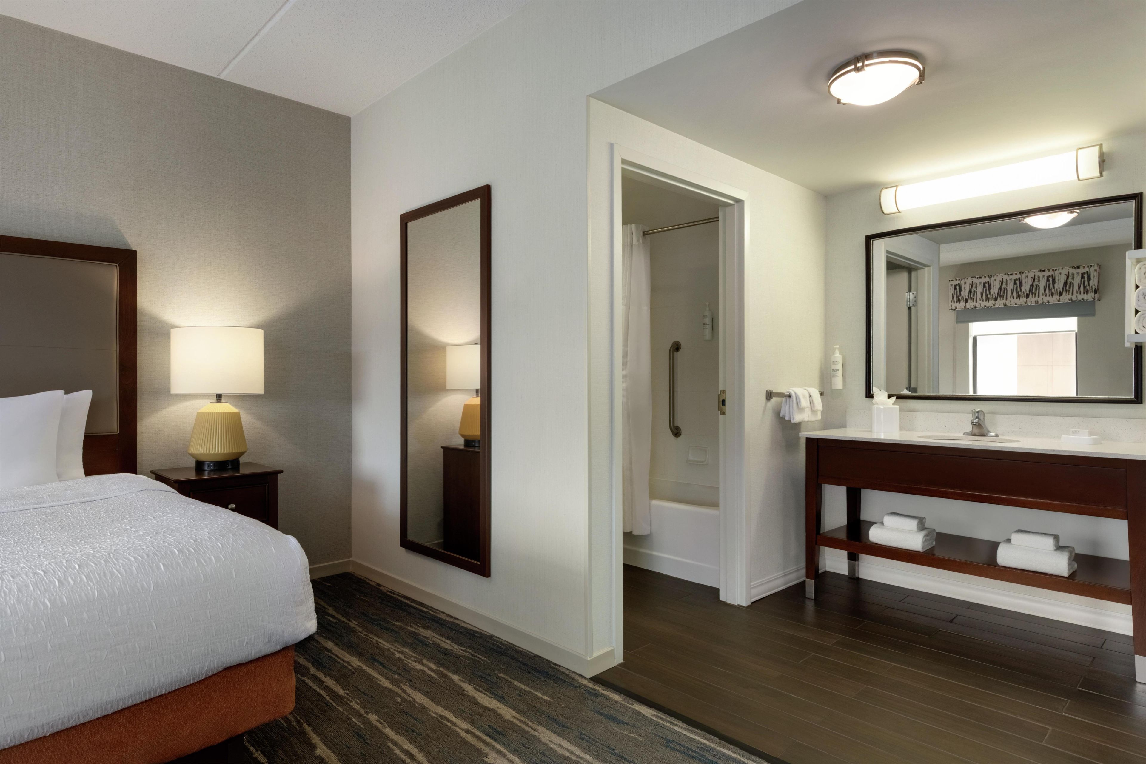 Habitación de hotel con cama, mesita de noche con lámpara, espejo grande, baño con ducha y tocador con espejo.