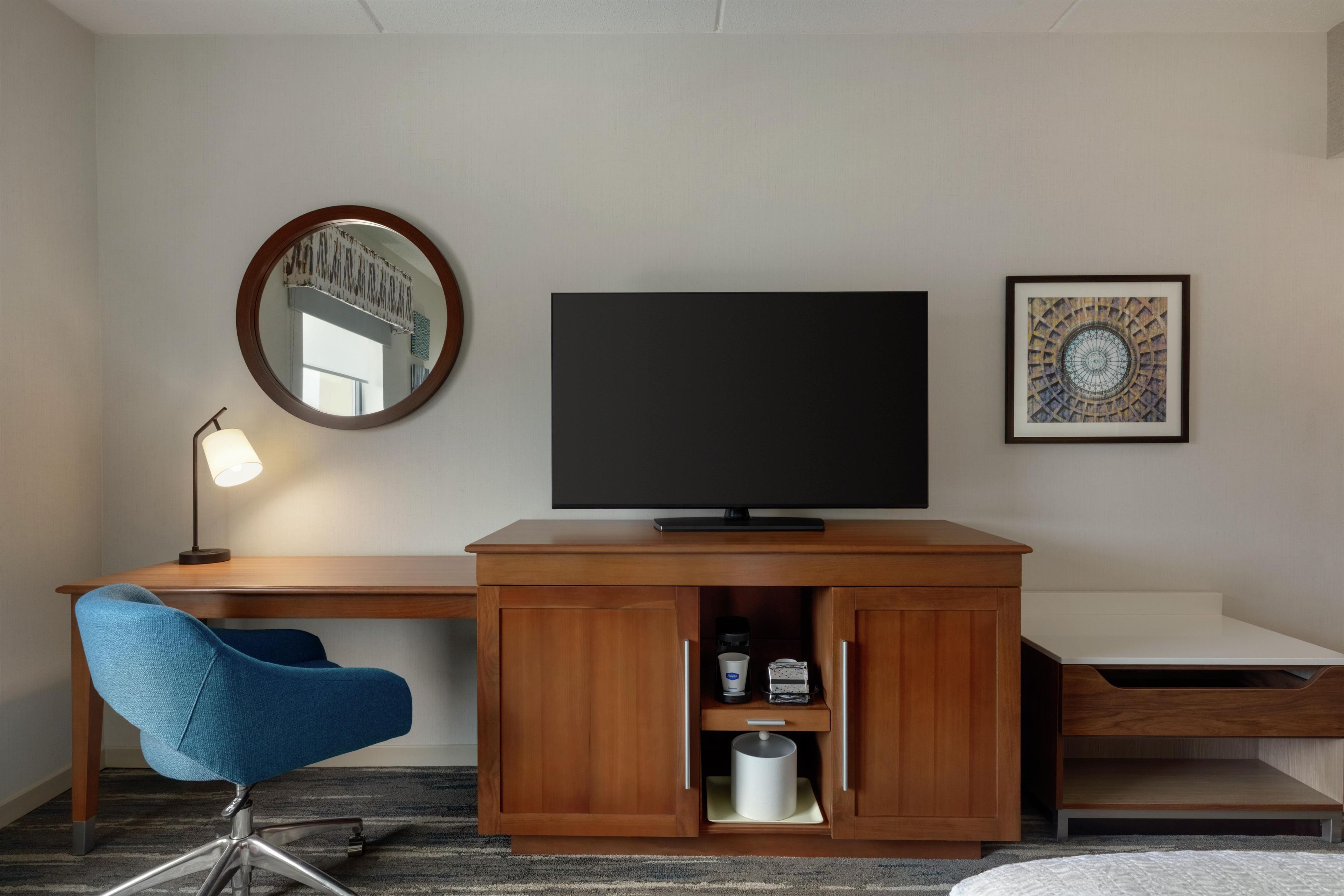 A hotel room with a wooden desk, a blue chair, a flat-screen TV, a round mirror, and a framed picture on the wall.