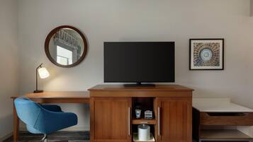 A hotel room with a wooden desk, a blue chair, a flat-screen TV, a round mirror, and a framed picture on the wall.