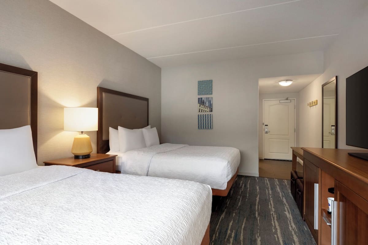 A hotel room with two beds, a desk, a lamp, a TV, and a door.