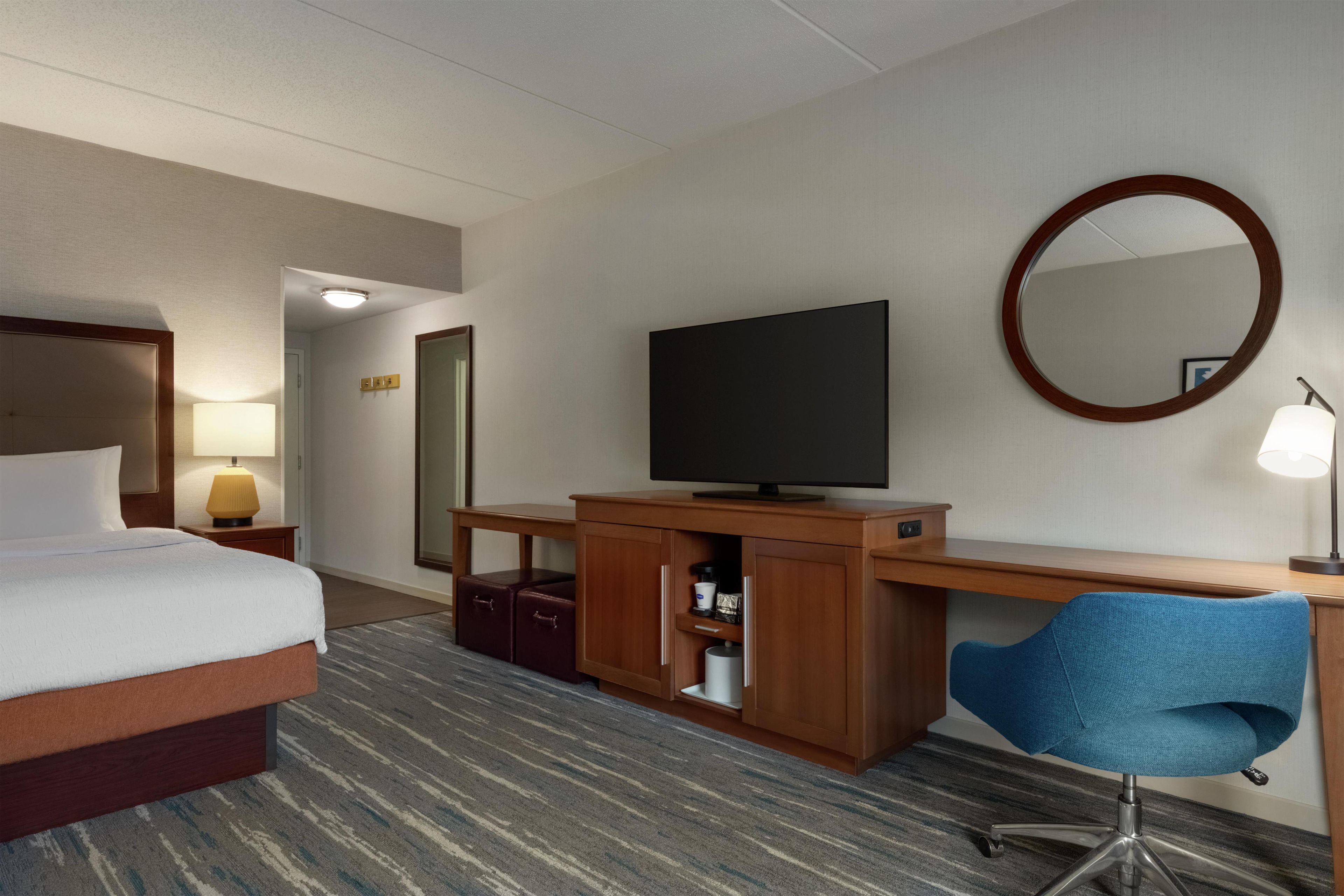 Quarto de hotel com cama, escrivaninha, cadeira, televisão e espelho.