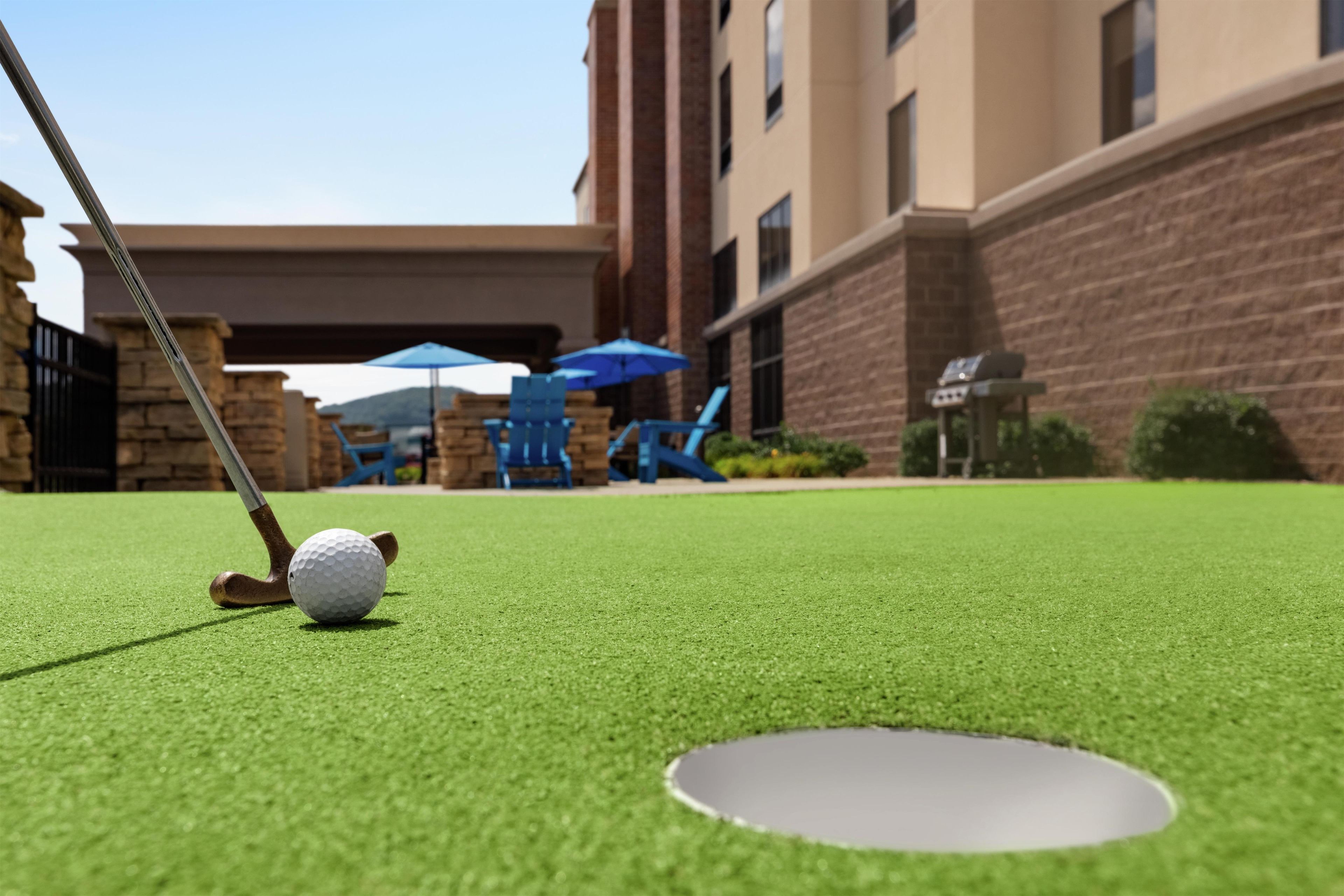 A golf ball on artificial turf near a hole, with a golf club in the foreground.