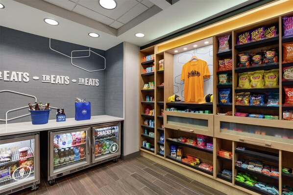 Un puesto de aperitivos con diversas botanas y bebidas empaquetadas, una camiseta colgando y un refrigerador con productos.