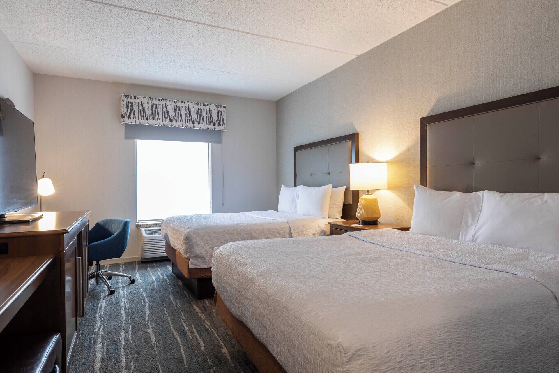 Habitación de hotel con dos camas, un escritorio, una silla y un ventanal grande.