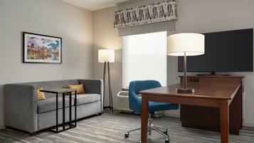 A hotel room with a grey sofa, a small table, a blue armchair, a desk with a lamp, a flat-screen TV, and a framed cityscape picture on the wall.