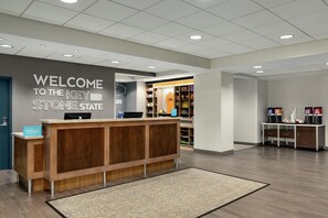 A hotel reception area with a wooden reception desk, a 'Welcome to the Key Stone State' sign, and a self-service kiosk.