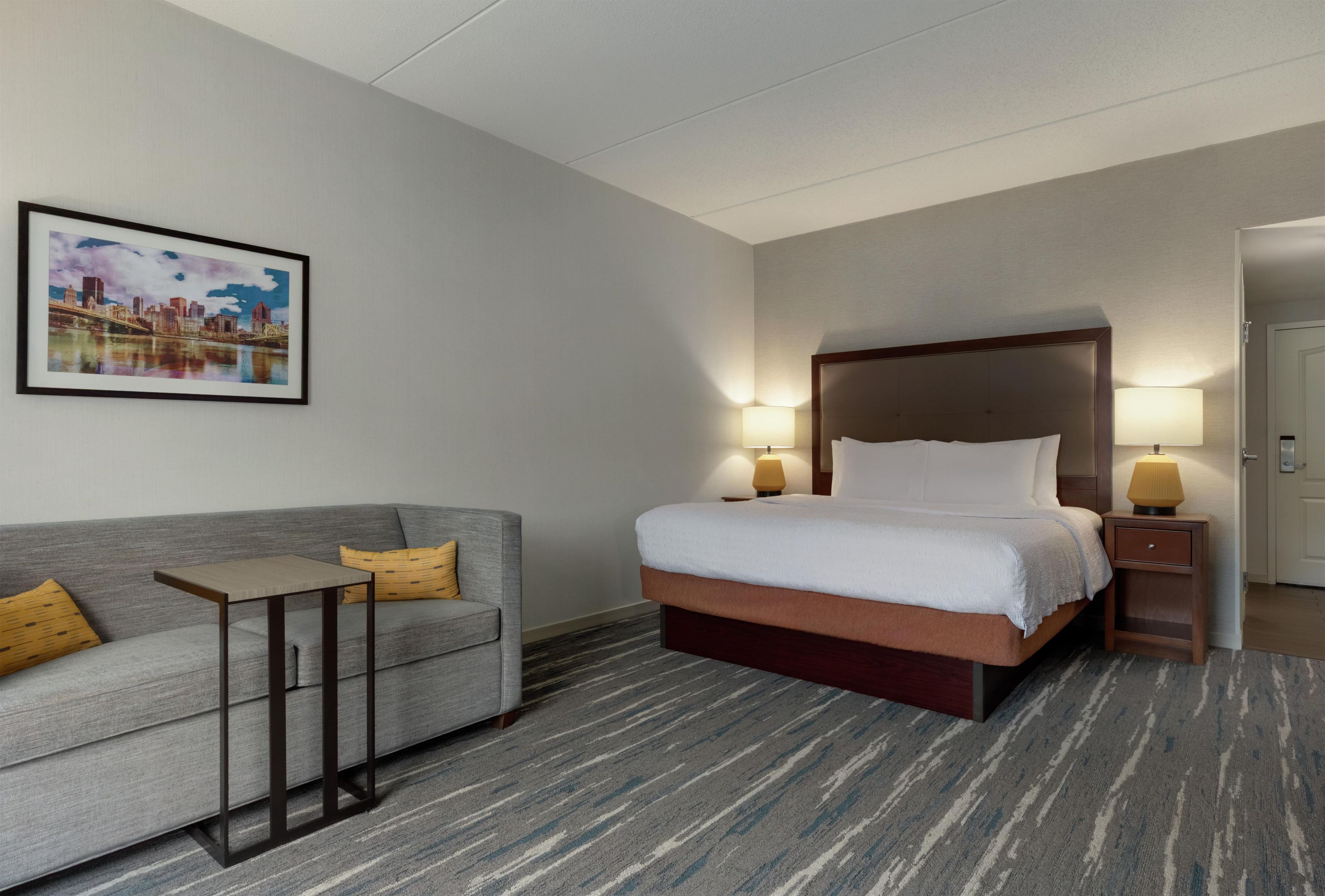 Habitación de hotel con cama, sofá, dos lámparas de noche y un cuadro de una vista urbana en la pared.