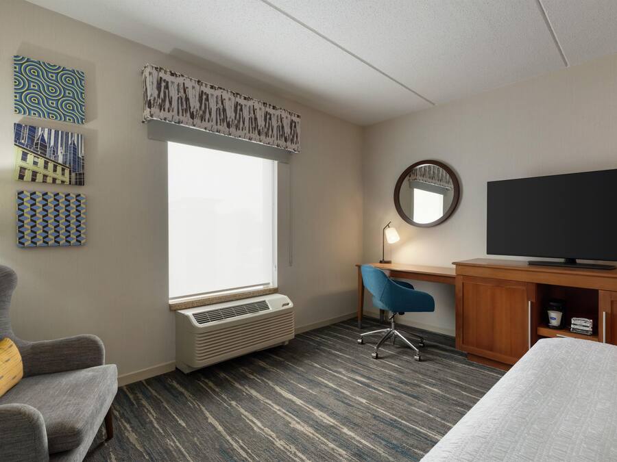 Una habitación de hotel con cama, escritorio con televisor, silla, calentador y obras de arte en las paredes.