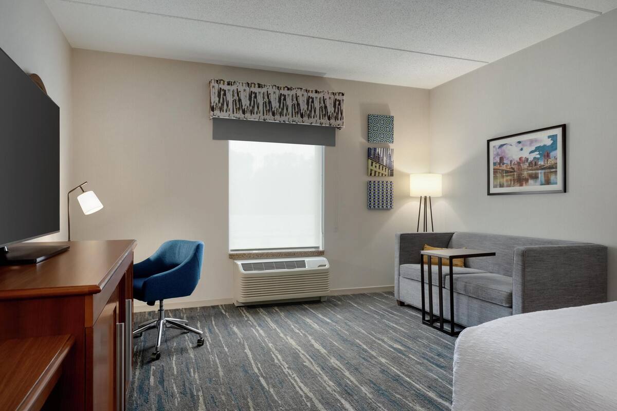 A hotel room with a desk, chair, bed, and a large window with an air conditioner.