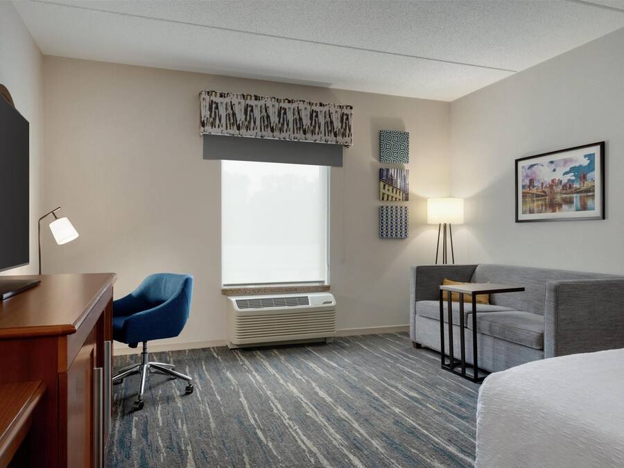A hotel room with a desk, chair, bed, and a large window with an air conditioner.