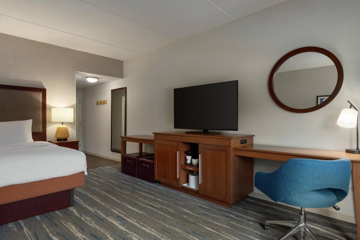 A hotel room with a bed, a desk with a computer, a television, a mirror, and a lamp.