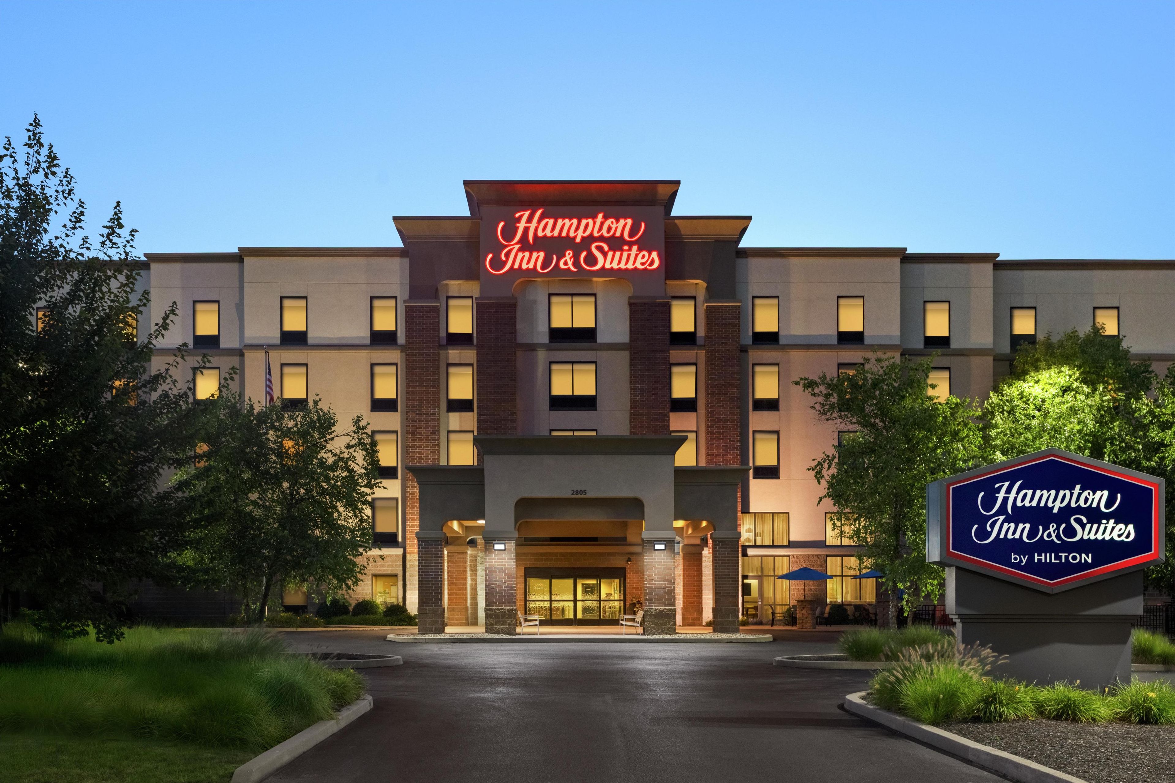 A Hampton Inn & Suites by Hilton building with a prominent sign, a well-lit entrance, and a landscaped driveway.