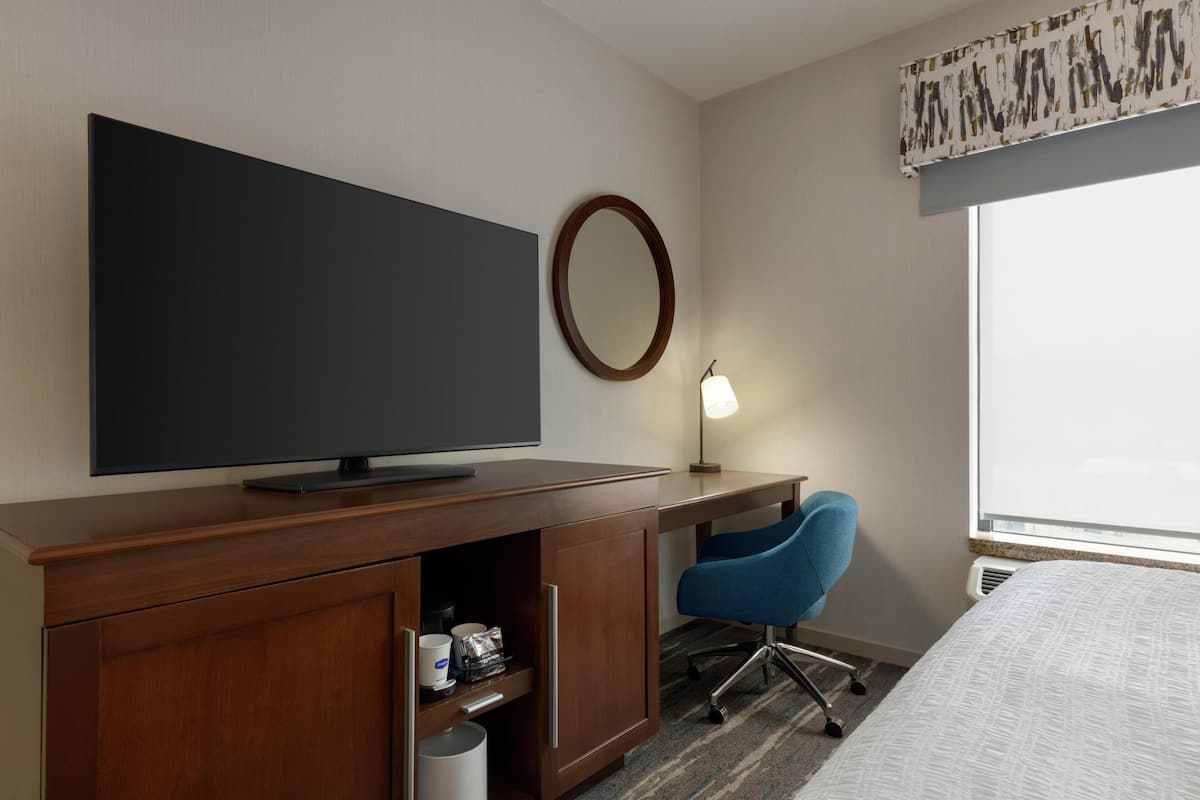 A hotel room with a flat-screen TV, a desk with a chair, a mirror, and a window with a view.