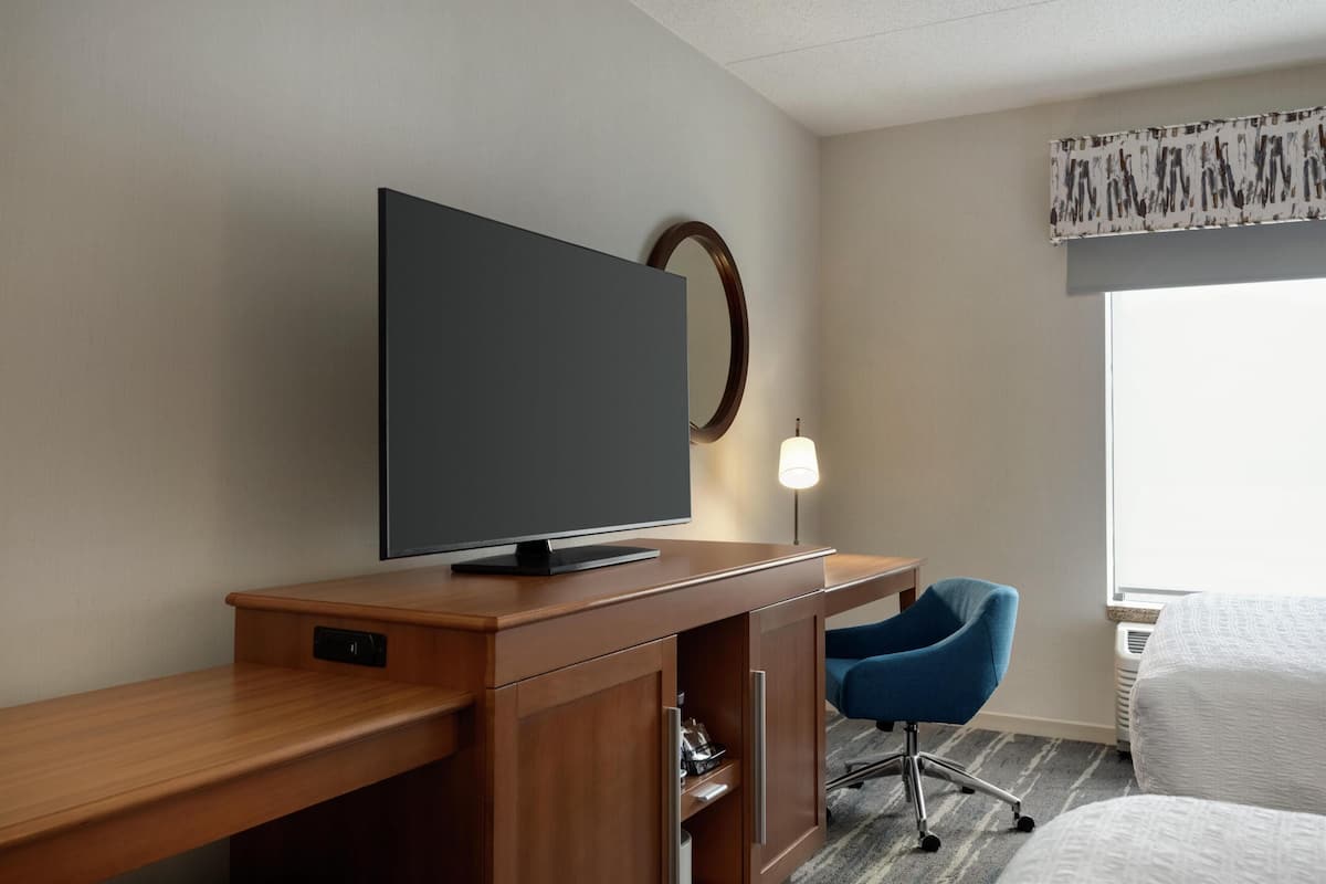 A hotel room with a wooden desk, a flat-screen TV, a blue chair, a bed, a window with a view, and a wall-mounted mirror.