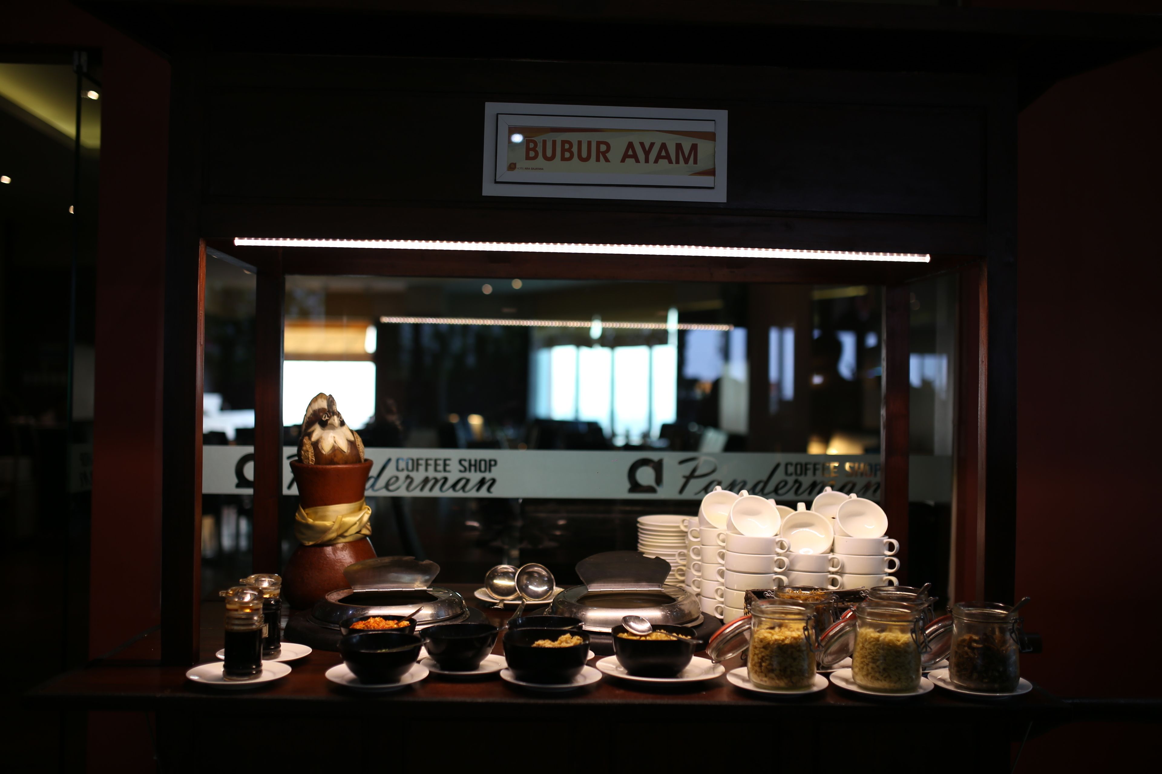 Un área de bufé con diversos alimentos y platos, un cartel que dice "BUBUR AYAM" y un letrero de cafetería al fondo.