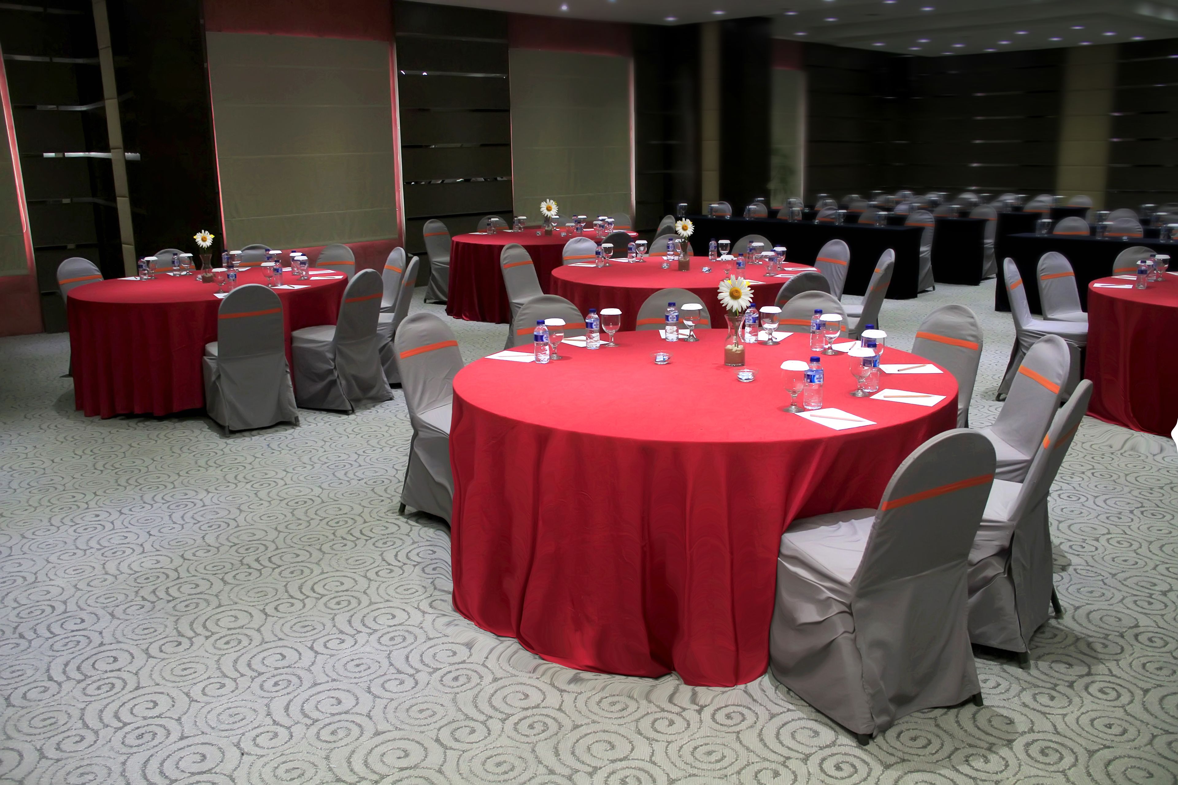 A conference room with multiple round tables covered with red tablecloths, each equipped with water bottles, glasses, and notepads.