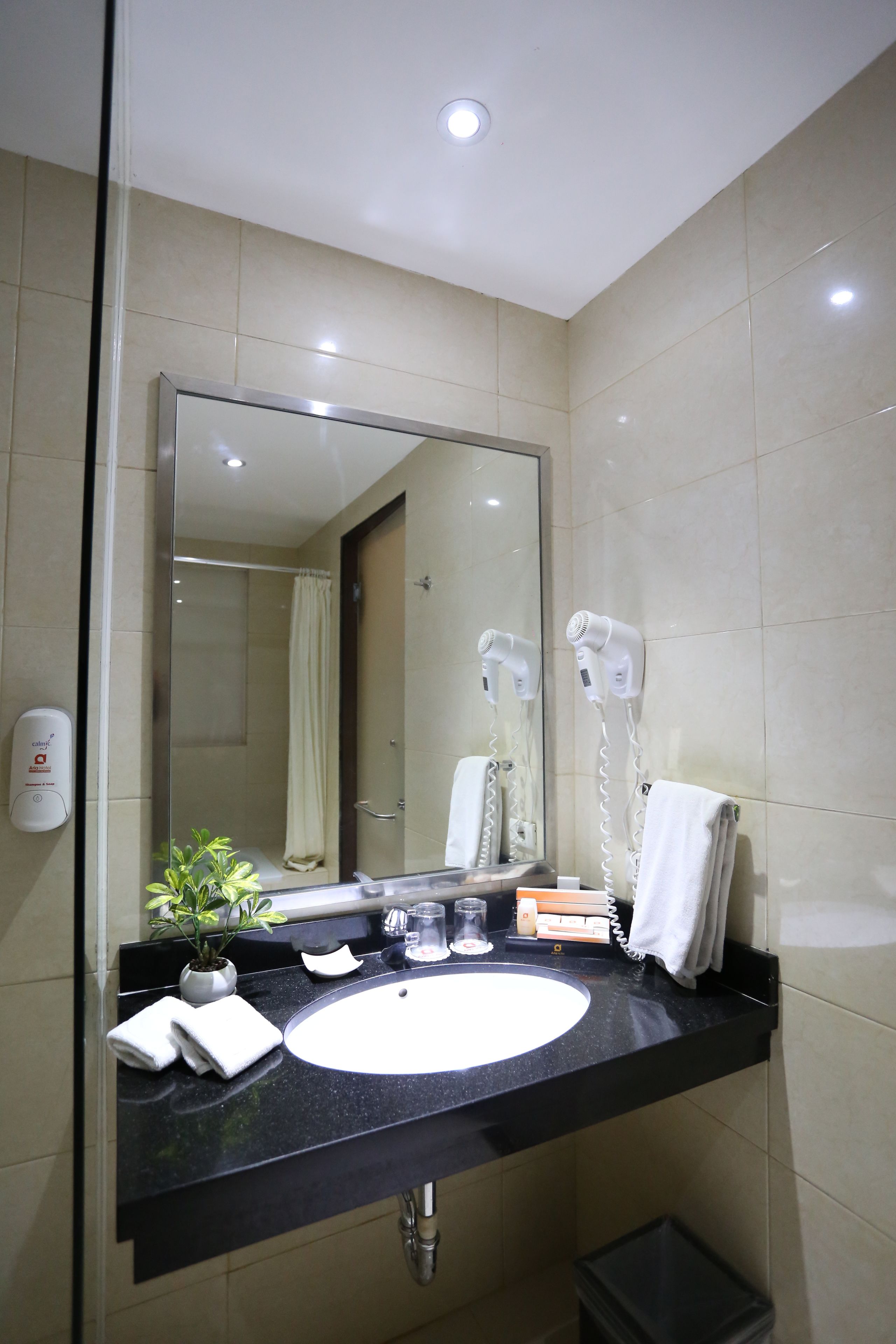 A bathroom with a large mirror, a sink, a hairdryer, a towel, and a trash bin.