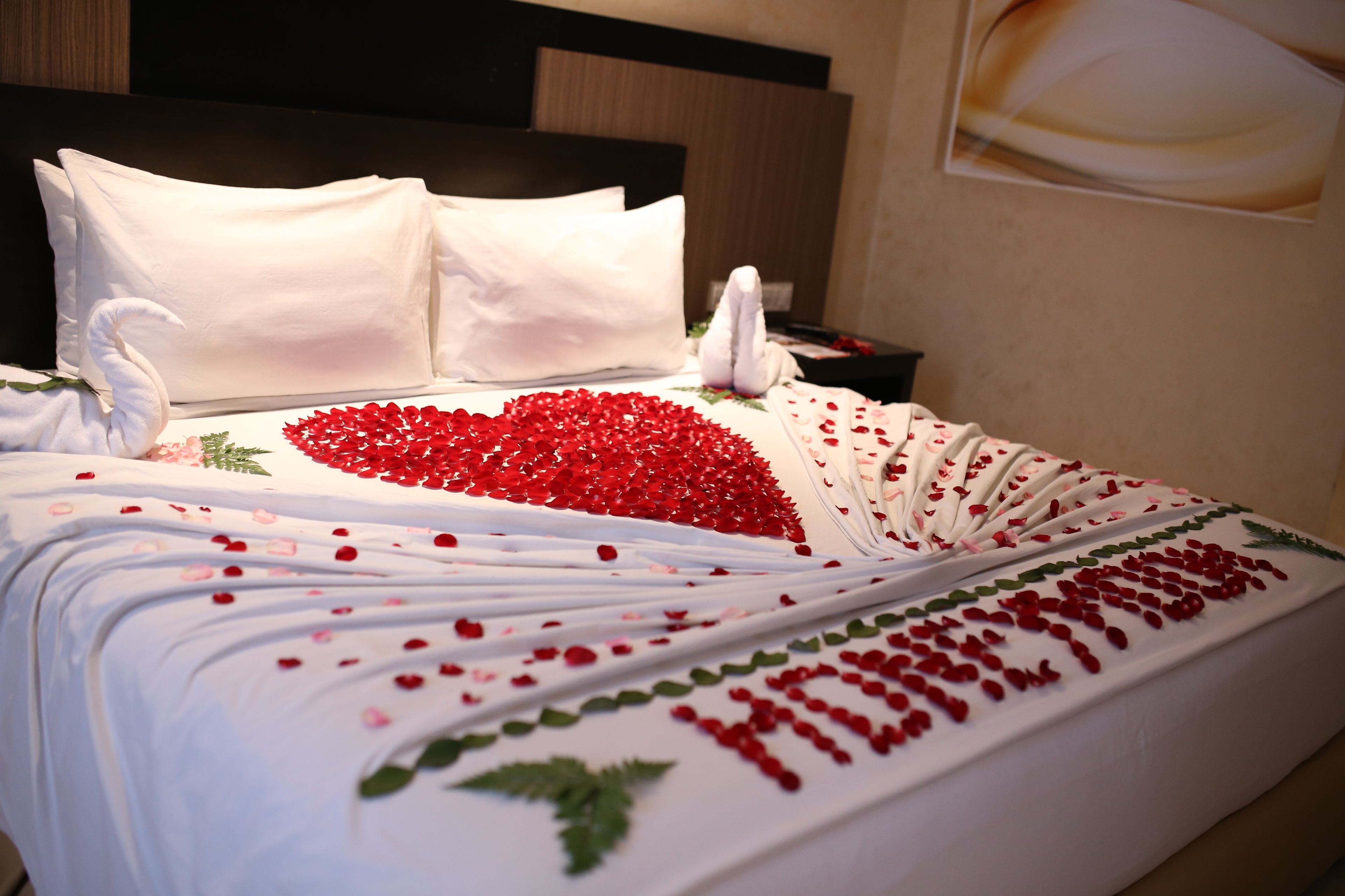 A bed with white linens, red rose petals, and a swan decoration.