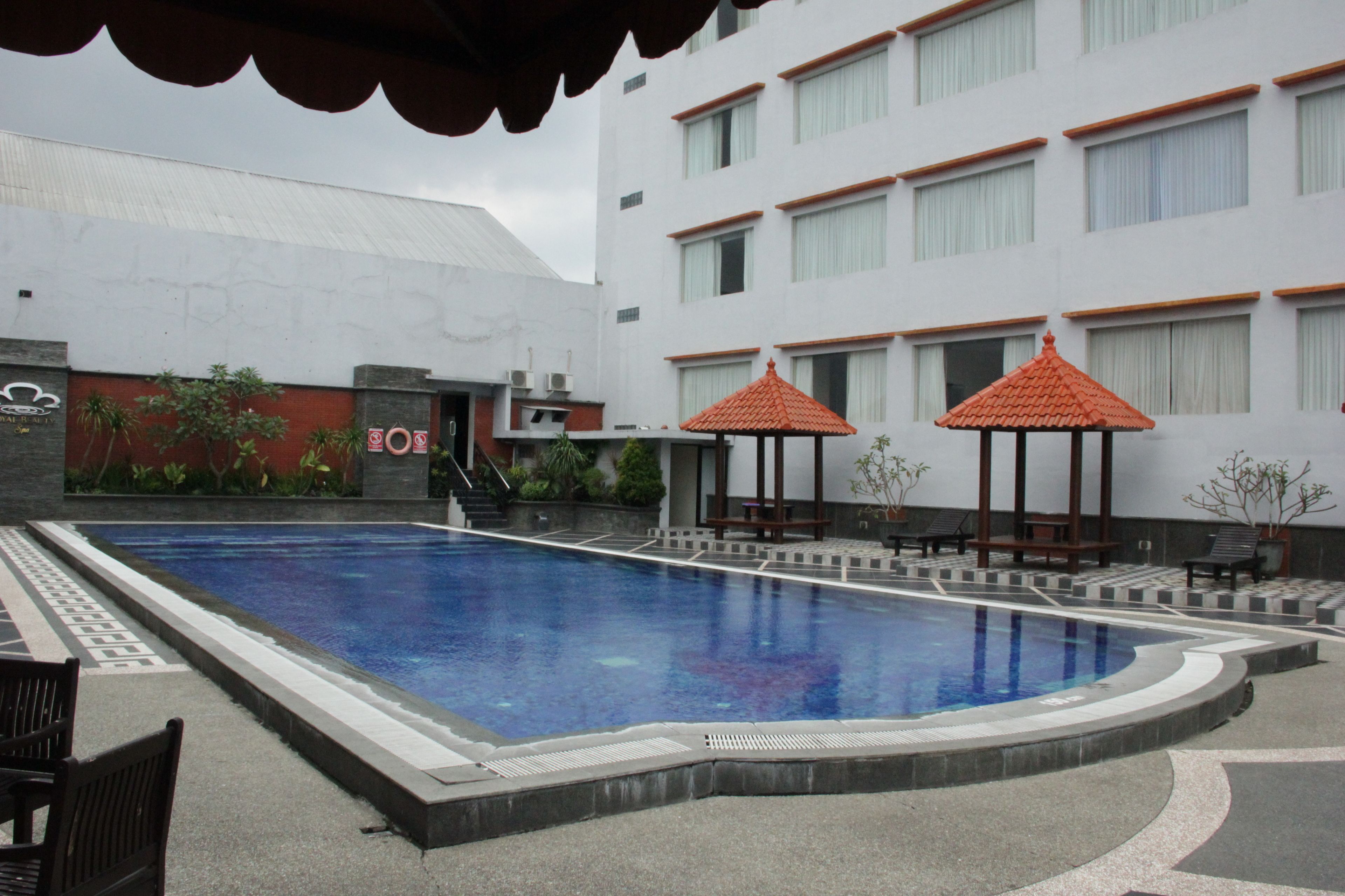A swimming pool with a tiled deck, surrounded by lounge chairs and umbrellas, and a building with multiple windows in the background.