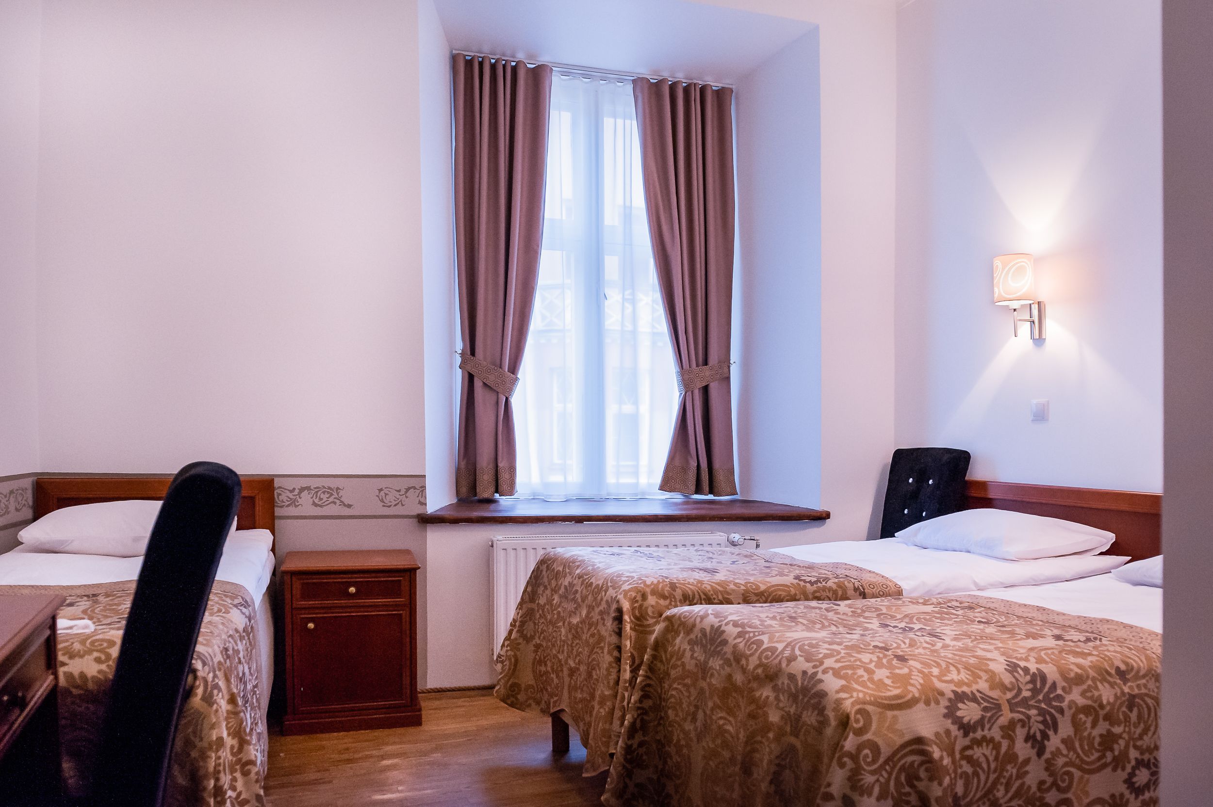A hotel room with two beds, a window with curtains, a bedside table, a chair, and a wall-mounted lamp.