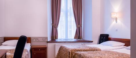 A hotel room with two beds, a window with curtains, a bedside table, a chair, and a wall-mounted lamp.