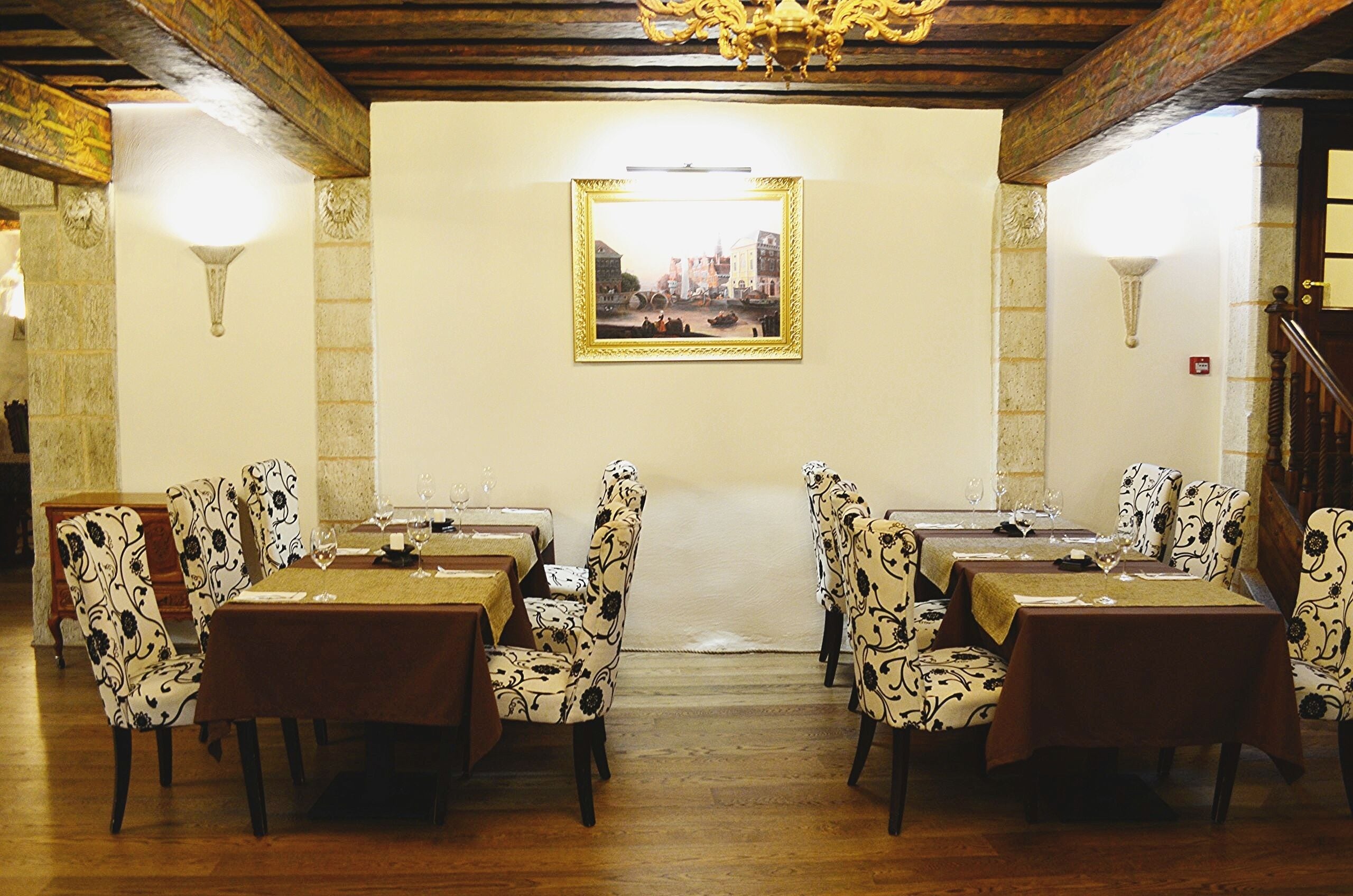 Un espace repas avec des tables dressées pour quatre personnes, chacune agrémentée d’un verre à vin et d’une assiette. La pièce est dotée de parquets en bois et d’un lustre.