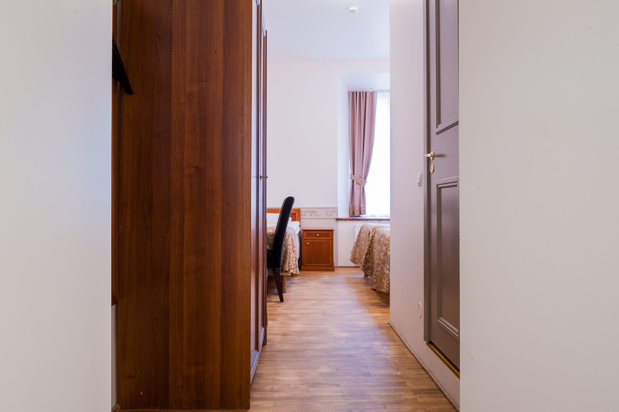 Un pasillo que conduce a un dormitorio con puerta de madera, cama con colcha estampada y ventana con cortinas.