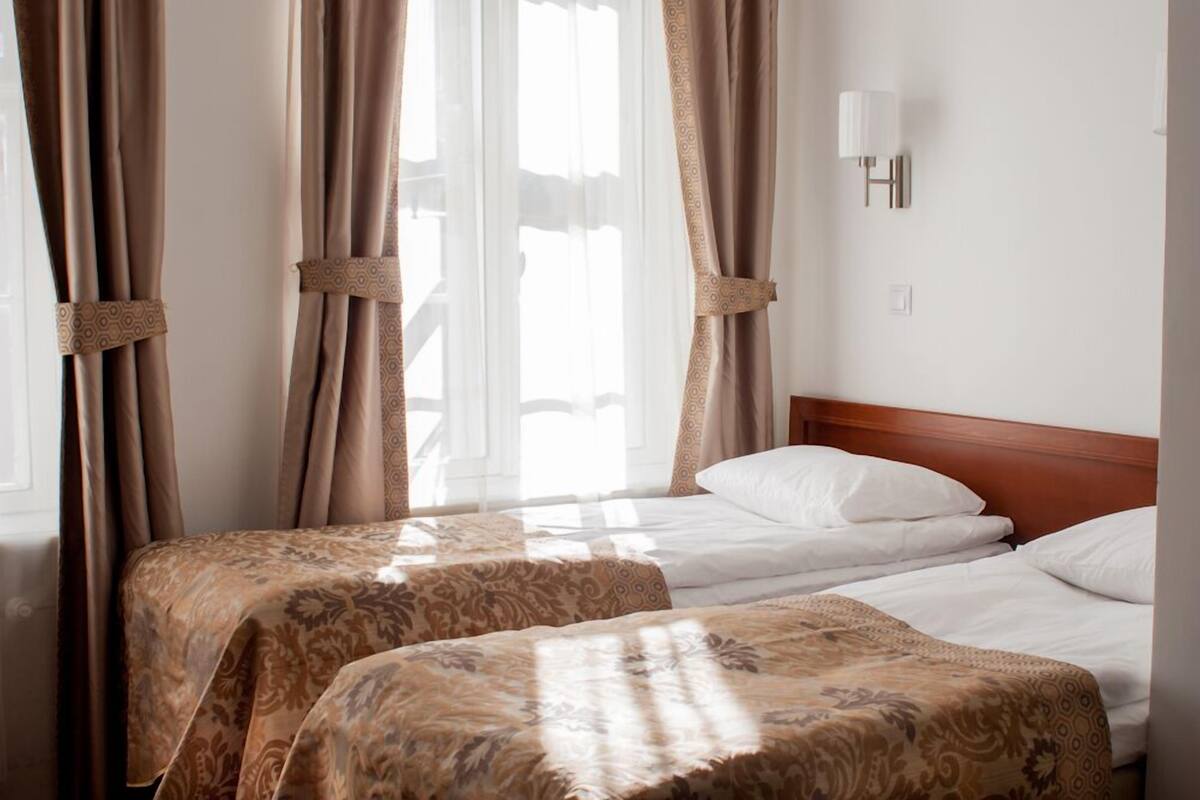 Ein Hotelzimmer mit zwei Betten, beigen Vorhängen, einem Holzkopfstück und einer wandmontierten Lampe.
