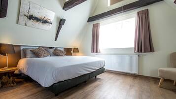 Un dormitorio con una cama, dos mesitas de noche, un ventanal con cortinas, un radiador y vigas de madera a la vista en el techo.