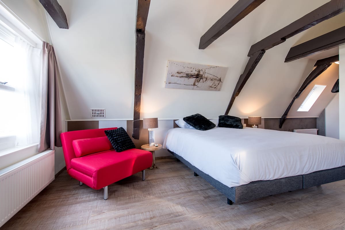 Ein Schlafzimmer mit rotem Sofa, einem großen Bett, Holzbalken, einem Fenster und einem Wandbild.