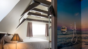 Un dormitorio con techo inclinado, una cama, una mesita de noche con lámpara, un ventanal con cortinas y un baño con ducha y espejo.