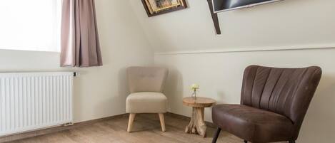 A room with a wooden floor, a beige armchair, a brown leather chair, a small wooden table with a vase, a framed picture, and a TV mounted on the wall.