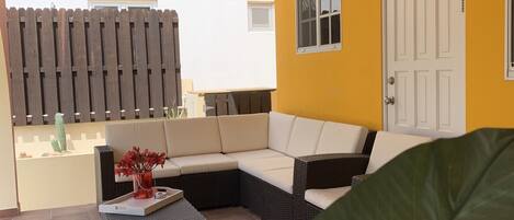 A covered outdoor seating area with a sofa and a table, a ceiling fan, and a door to the right.