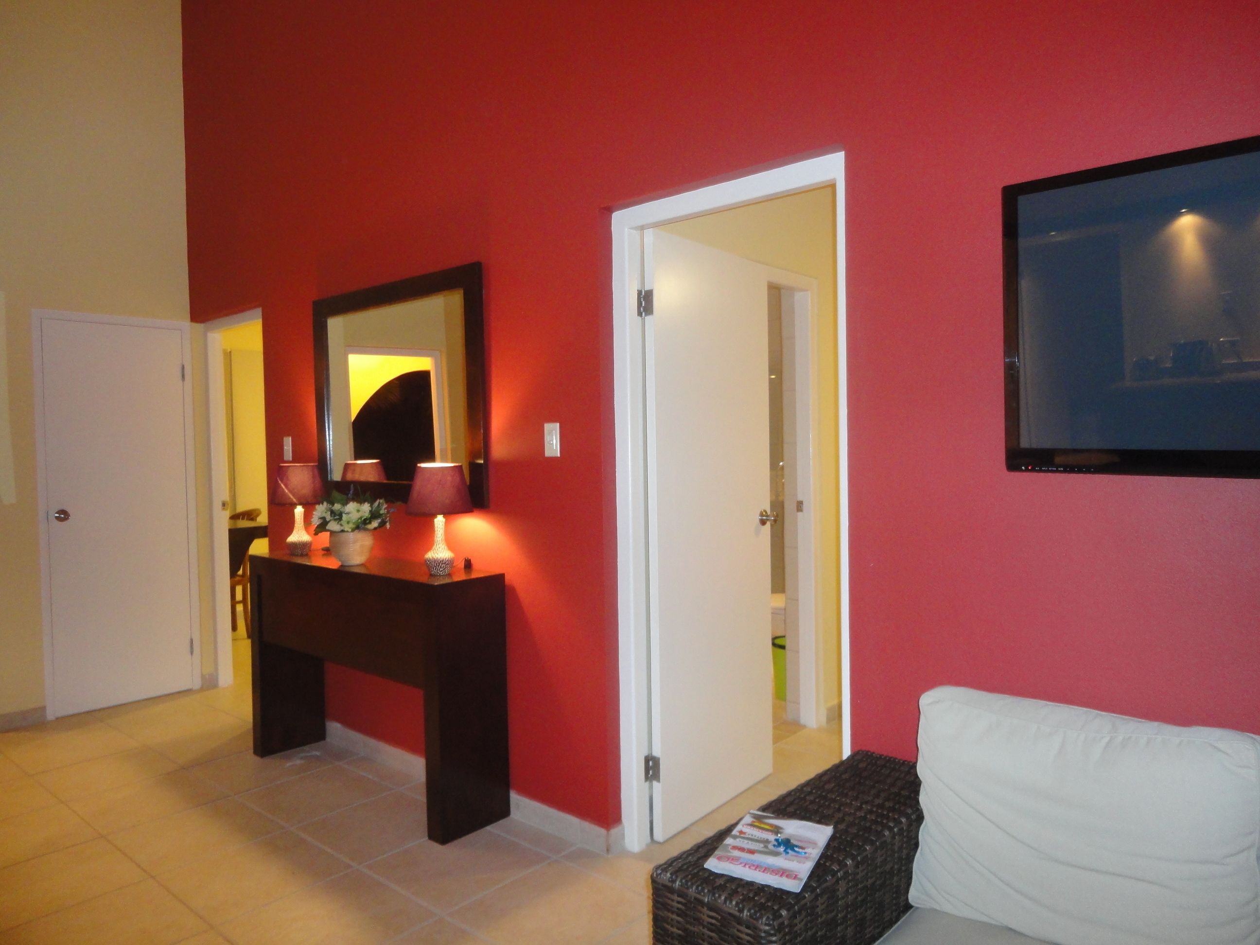 Ein Hotelzimmer mit roter Wand, einer weißen Tür, einem Holzkonsolentisch mit Lampen, einem weißen Sofa und einem an der Wand befestigten Flachbildfernseher.