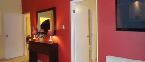 Ein Hotelzimmer mit roter Wand, einer weißen Tür, einem Holzkonsolentisch mit Lampen, einem weißen Sofa und einem an der Wand befestigten Flachbildfernseher.