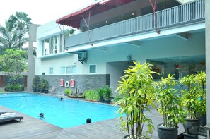 A swimming pool with a wooden deck, potted plants, and a building with a balcony.