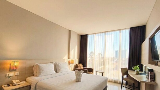 A hotel room with a bed, bedside table, lamp, chair, desk, and a large window. - Grand Cakra Hotel (Malang)