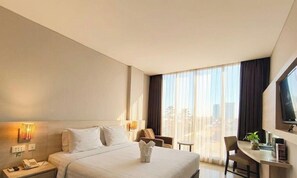 A hotel room with a bed, bedside table, lamp, chair, desk, and a large window. - Grand Cakra Hotel (Malang)