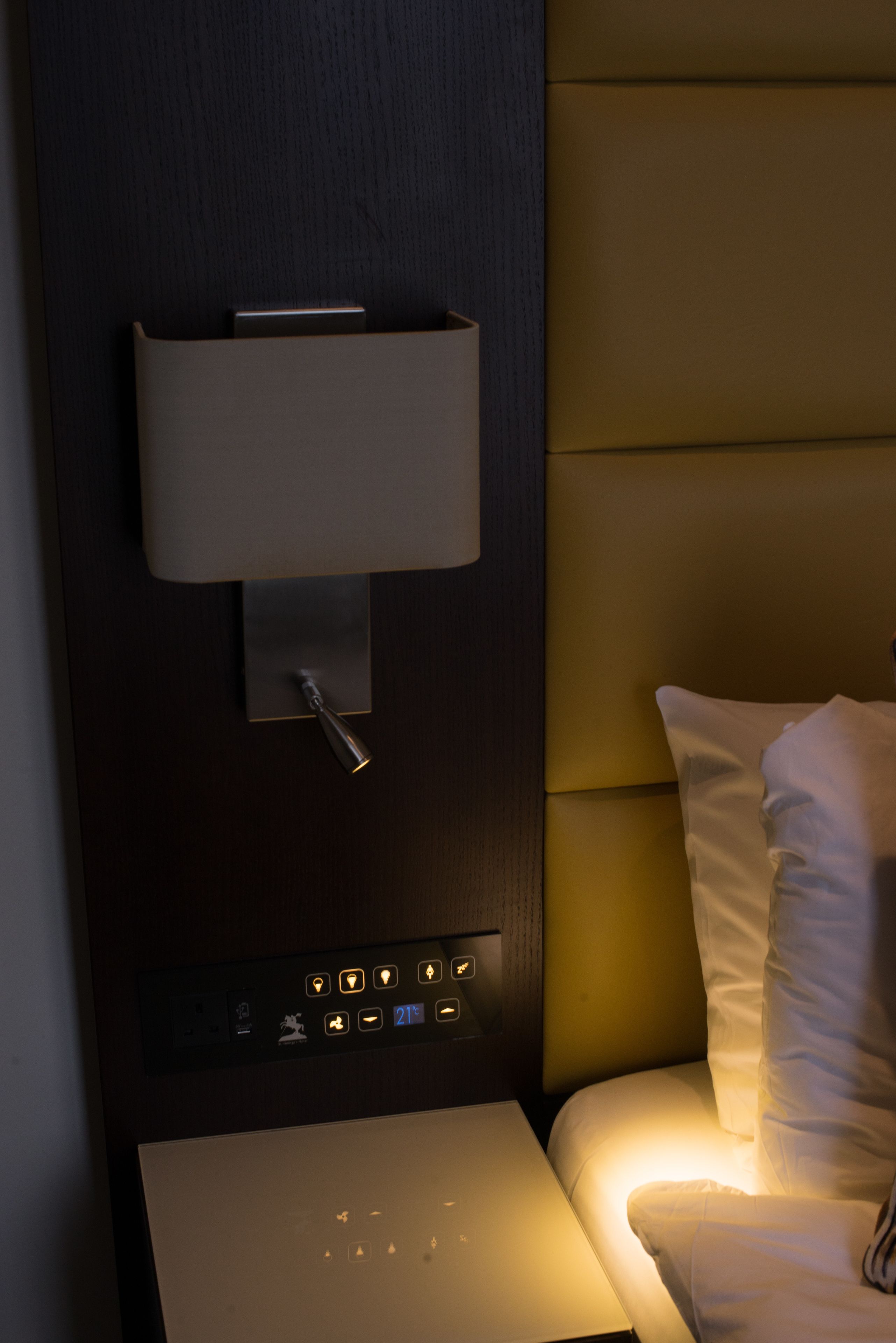 A modern hotel room with a dark wood headboard, a bedside table with a lamp, and a control panel with illuminated buttons.