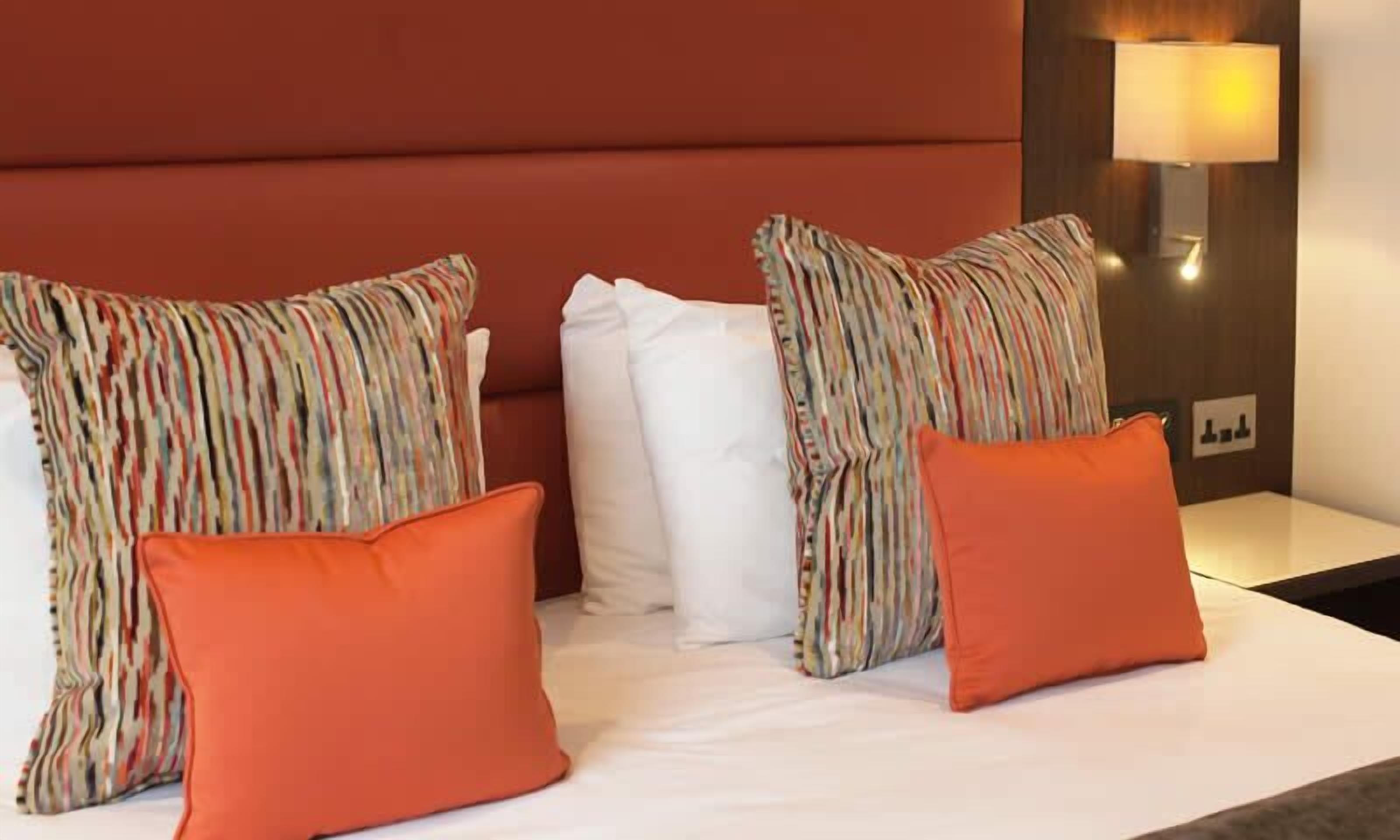 A hotel room with a bed, striped pillows, a white pillow, and an orange pillow.